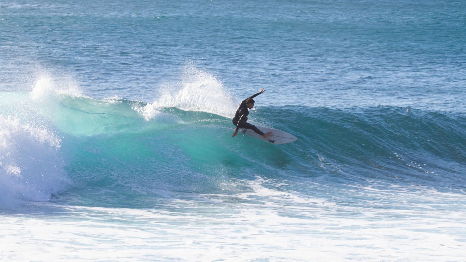 Surfer in black wetsuit riding a long turquoise wave with white foam on a bright sunny day.