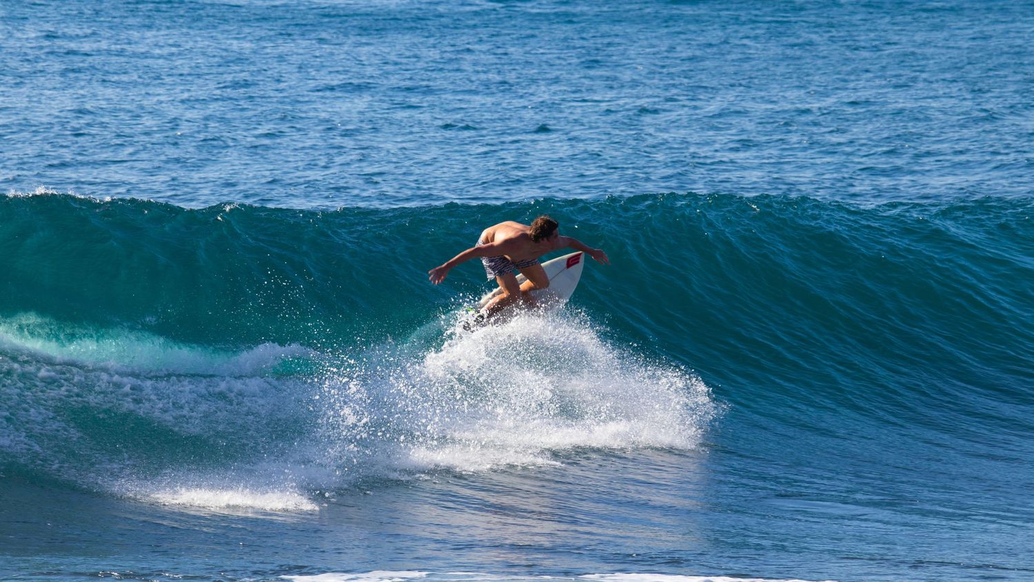 Surfer riding a turquoise wave without a wetsuit in clear tropical waters.