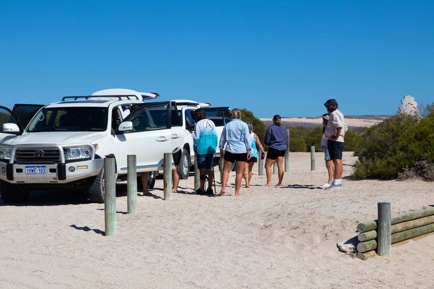 Group of tourists standing beside white 4WD vehicles on a sandy coastal Australian landscape.