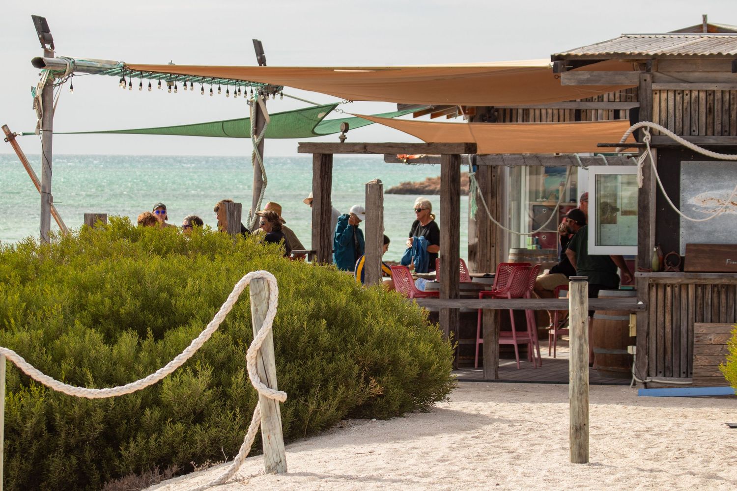 Rustic beachside bar with shade sails and wooden structure, patrons enjoying drinks overlooking turquoise ocean waters.