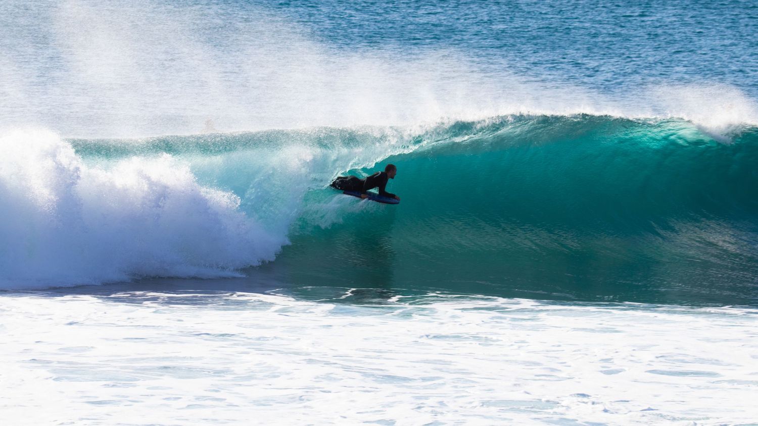 Surfer crouching deep inside a massive turquoise barrel wave with white water crashing overhead.