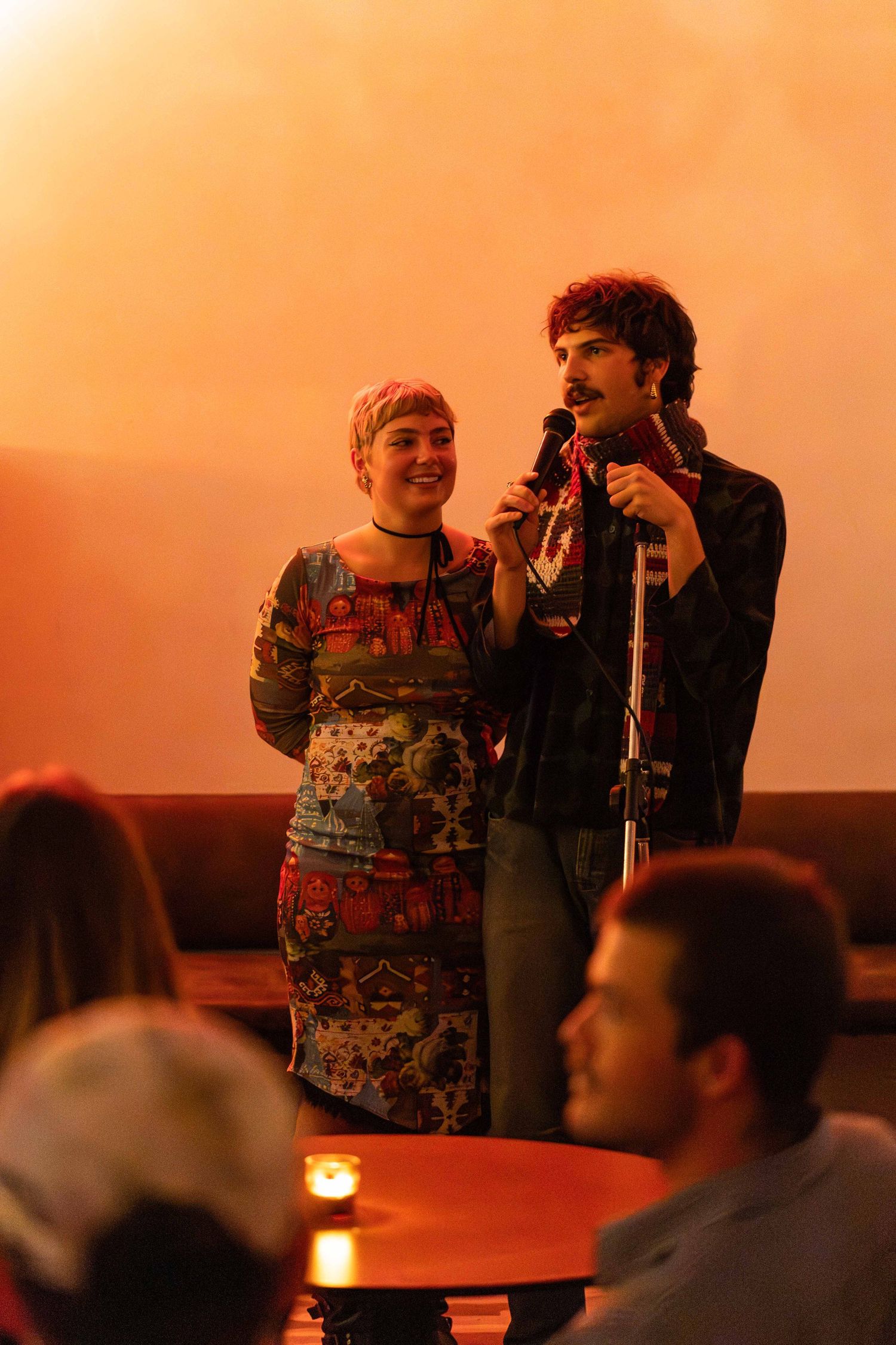 A man and woman share a microphone at a cozy, warmly lit venue event.