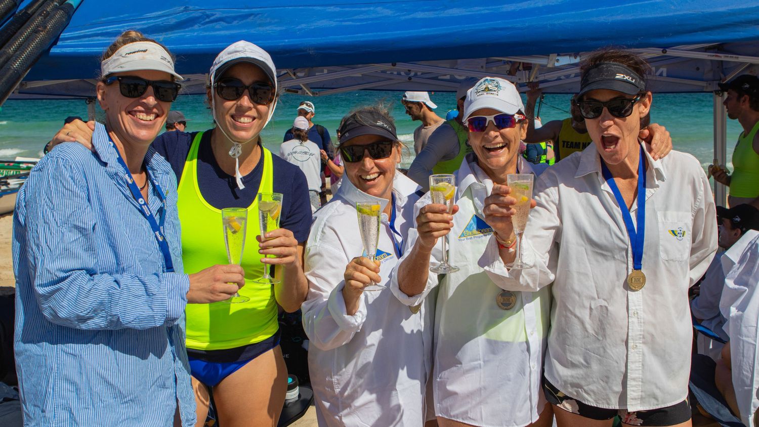 Five women celebrating with champagne glasses at a beach surf lifesaving event on a sunny day.