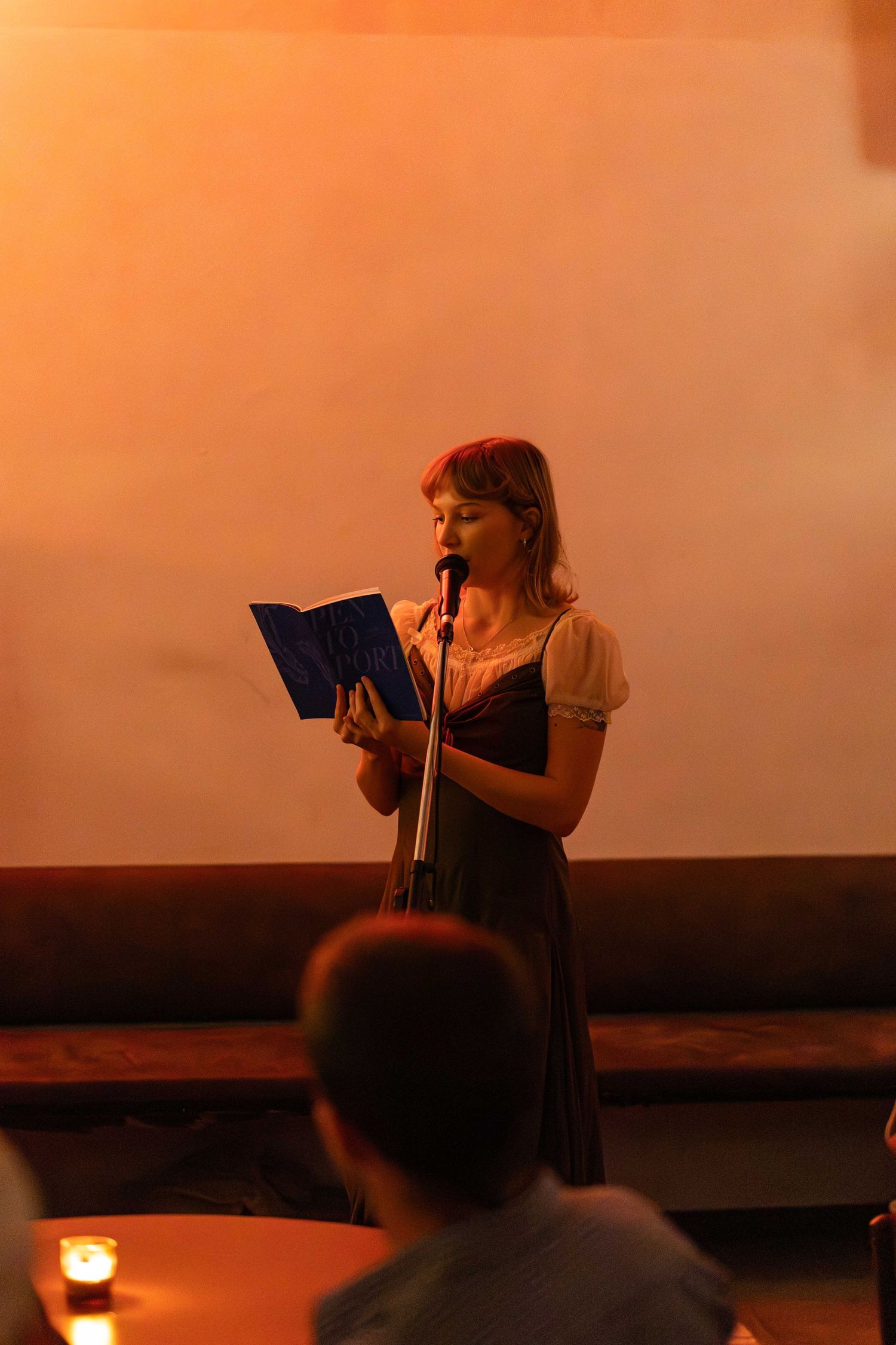A young woman reads from a blue book at a microphone stand in a warmly lit venue.