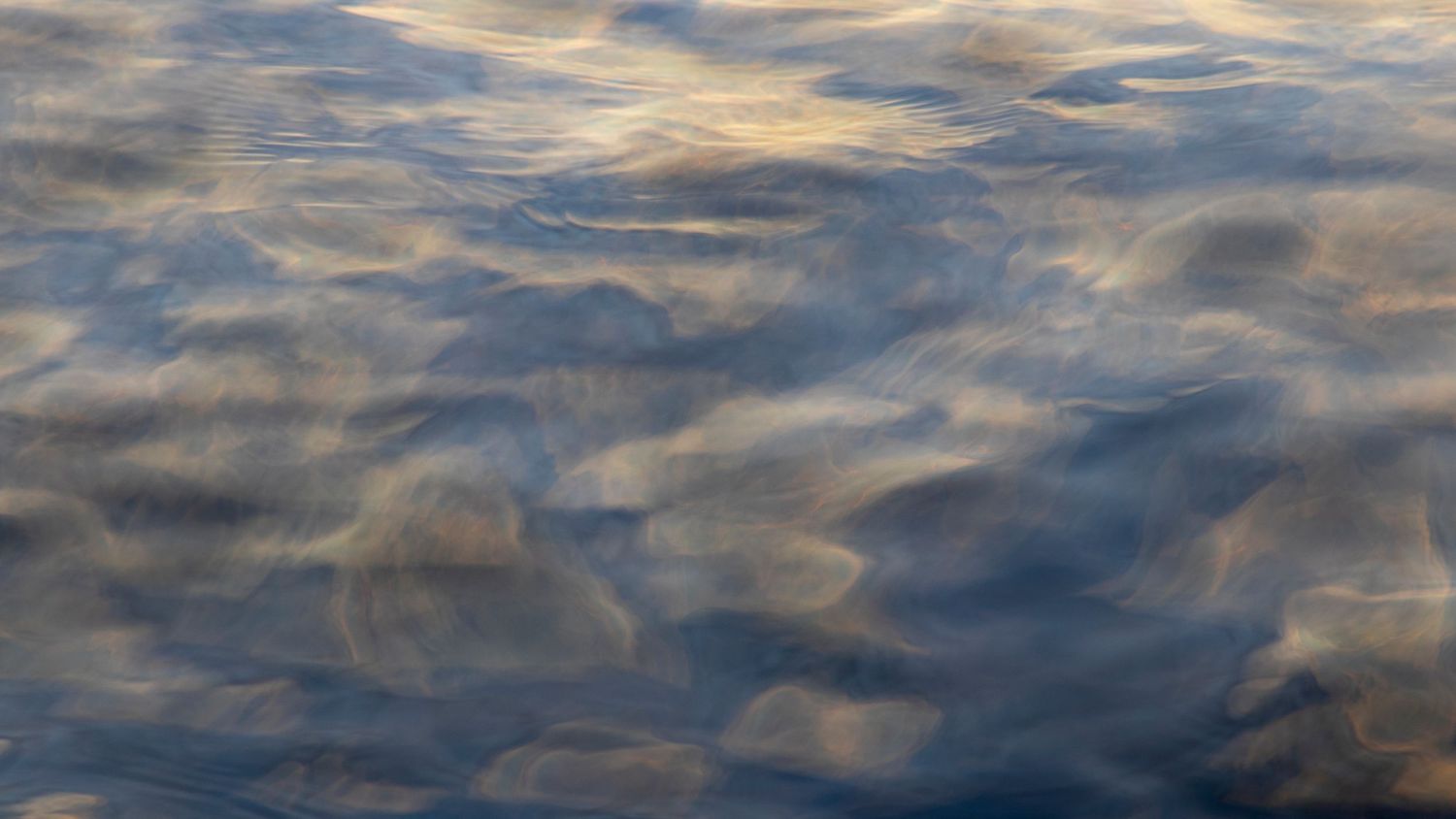 Calm blue water surface reflecting soft clouds with gentle ripples creating abstract patterns.