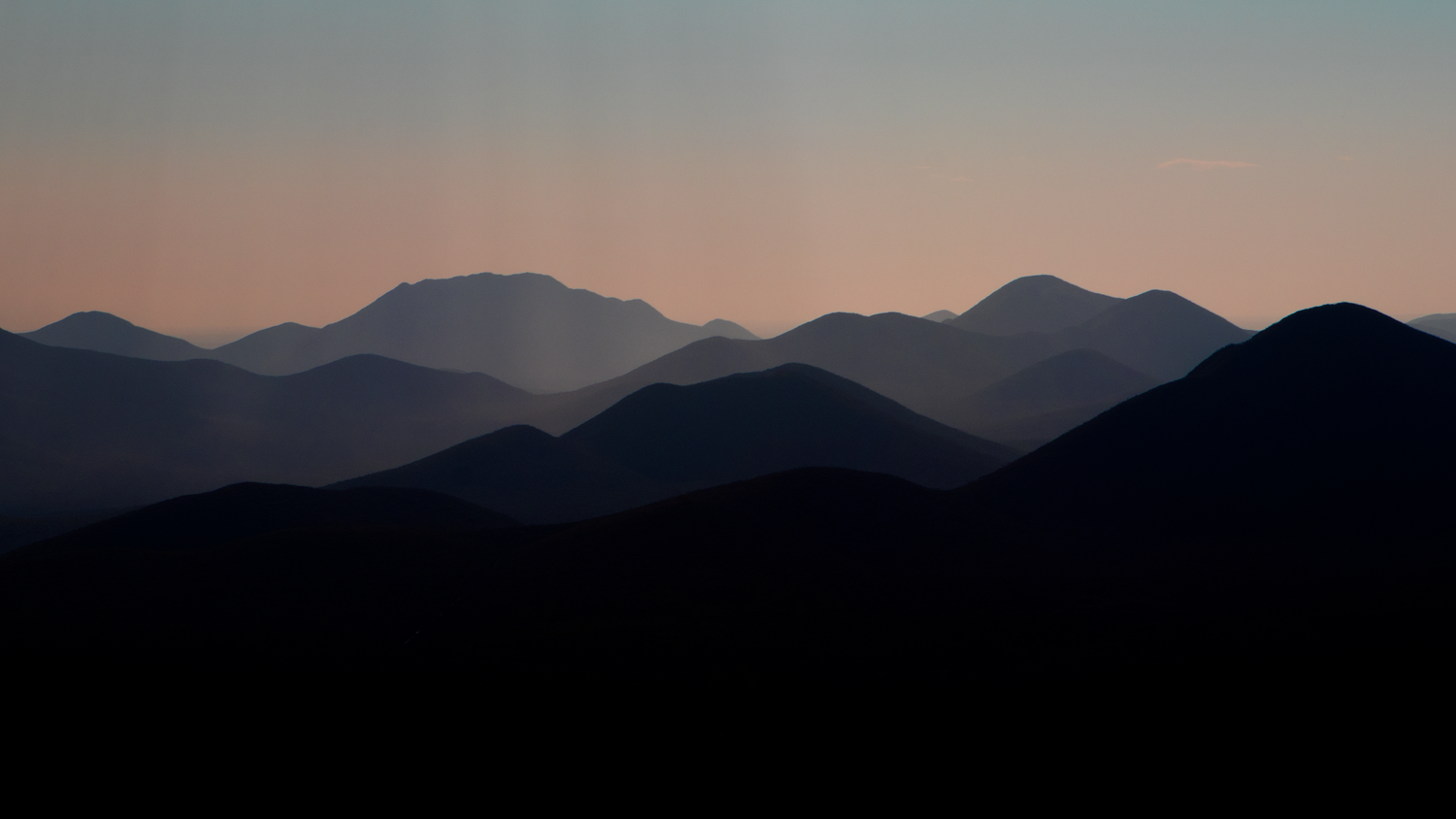 Silhouetted mountain ridges layered in blue haze against a soft pink and blue twilight sky at dusk.