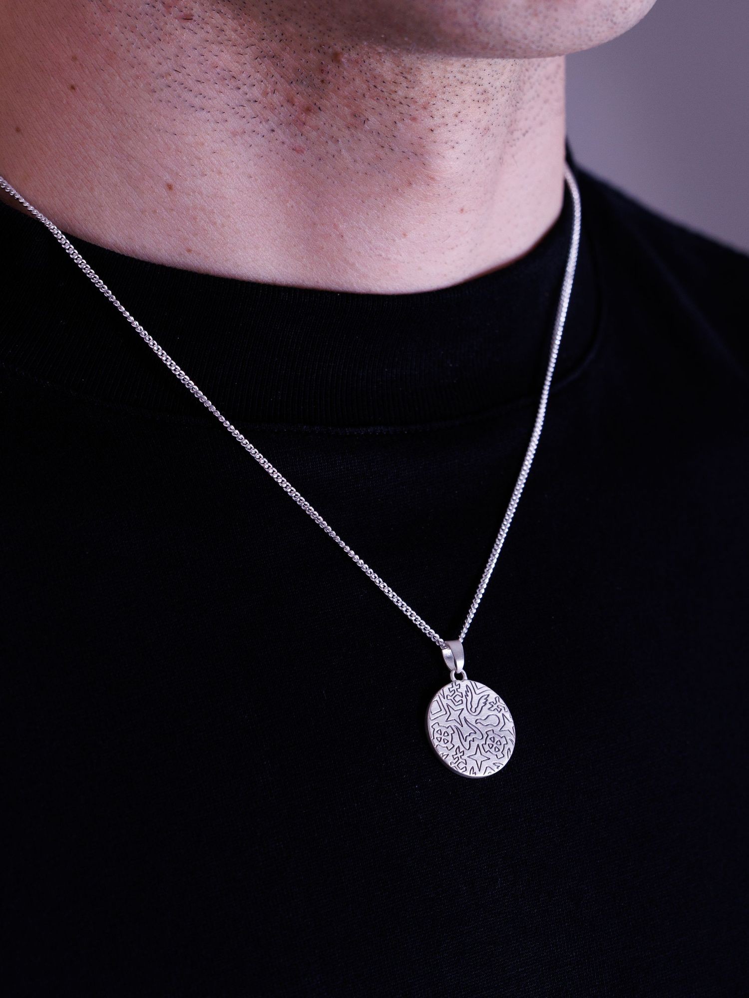 Man wearing a silver circular pendant necklace with intricate engraved pattern on a box chain.