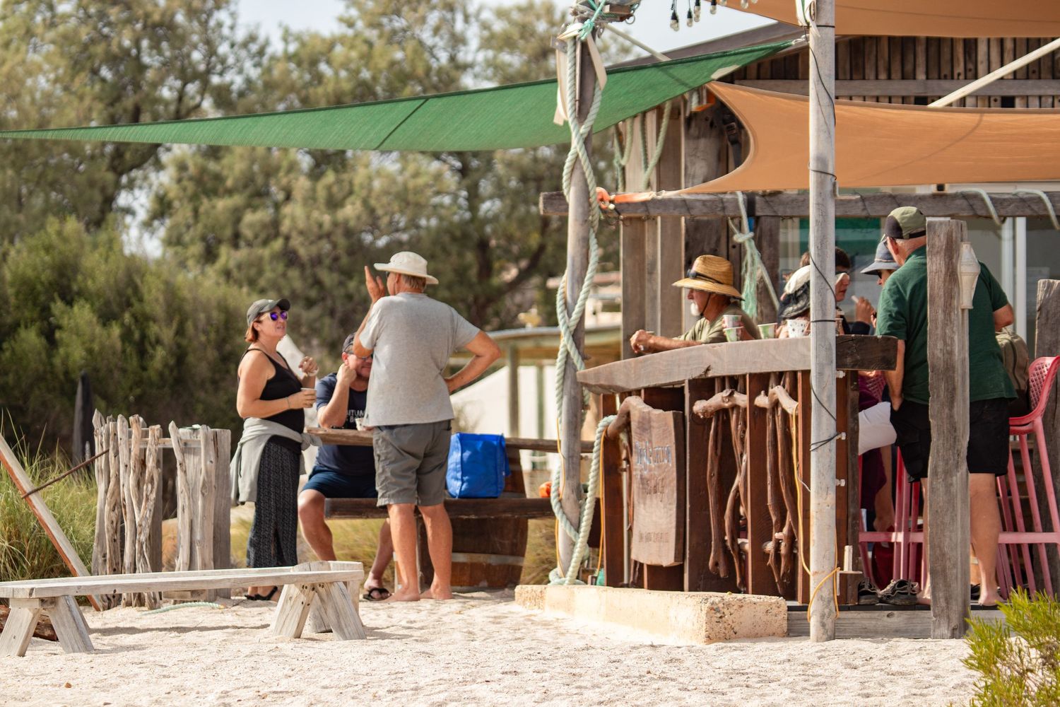Visitors ordering drinks at a rustic outdoor beach bar with green shade sails on a sunny day.