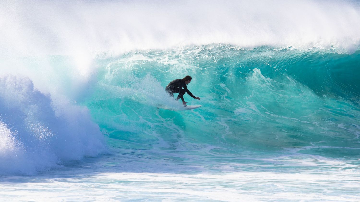 A surfer drops down the face of a massive turquoise barreling wave in open ocean.