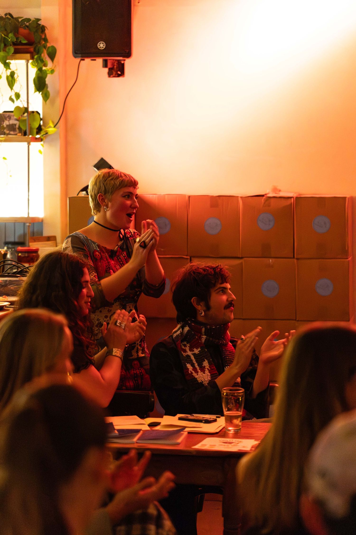 Audience members clapping and standing at a warmly lit indoor event with cardboard boxes stacked against the wall.