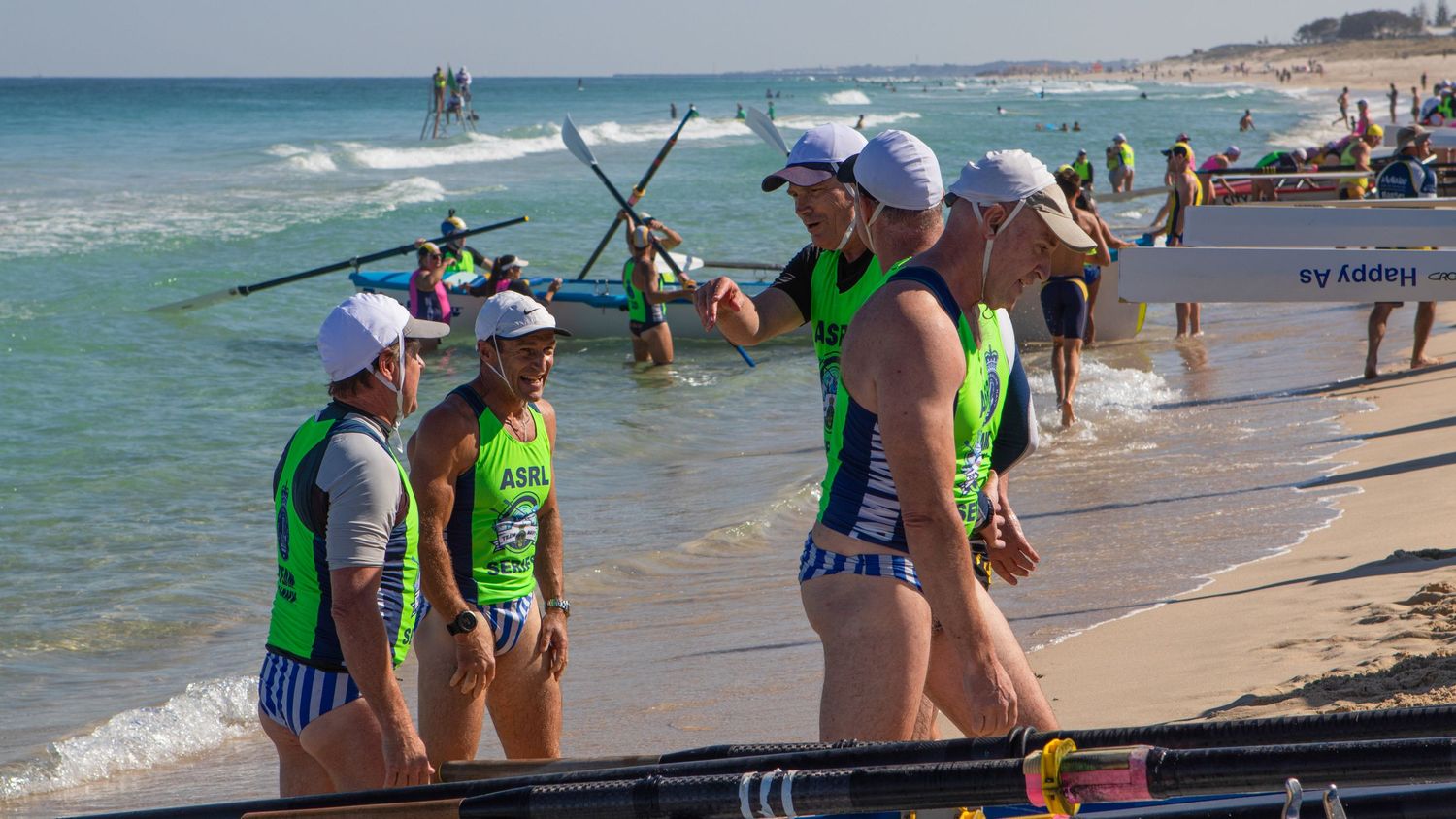 Surf boat rowing crew in ASRL Series vests carry their boat along an Australian beach shoreline.