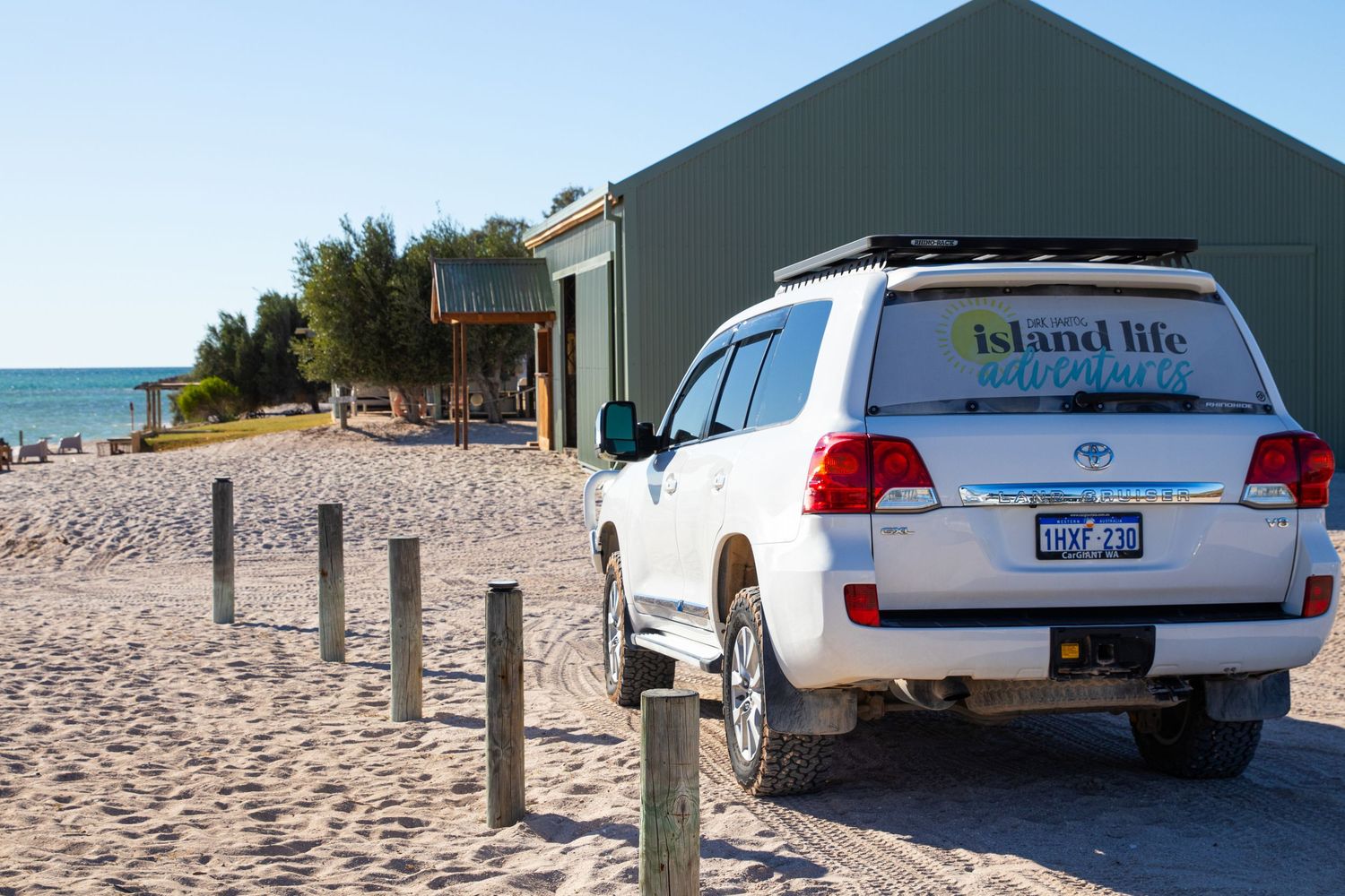 White Island Life Tours 4WD Toyota parked on beach sand beside green coastal building on sunny day.
