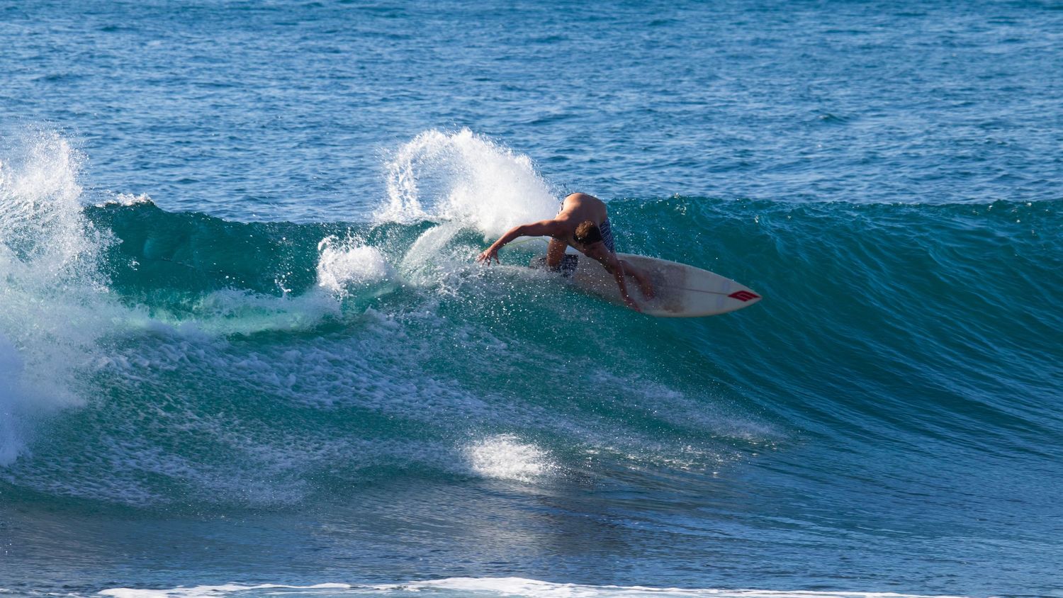 Surfer in red shorts performing a sharp turn on a white surfboard over a turquoise ocean wave.
