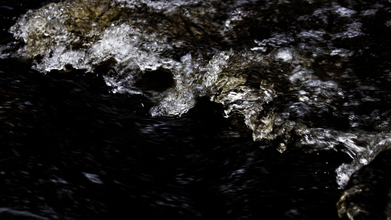 Dark rushing water cascades over rocks, creating white foam and splashes in a dramatic close-up shot.