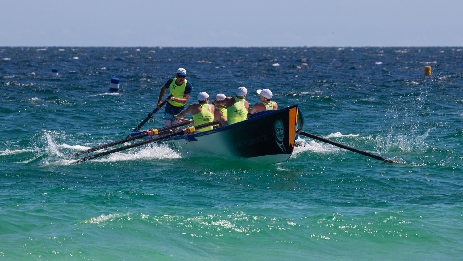 Surf boat crew in high-visibility yellow uniforms navigating dark hull through ocean waves competitively.