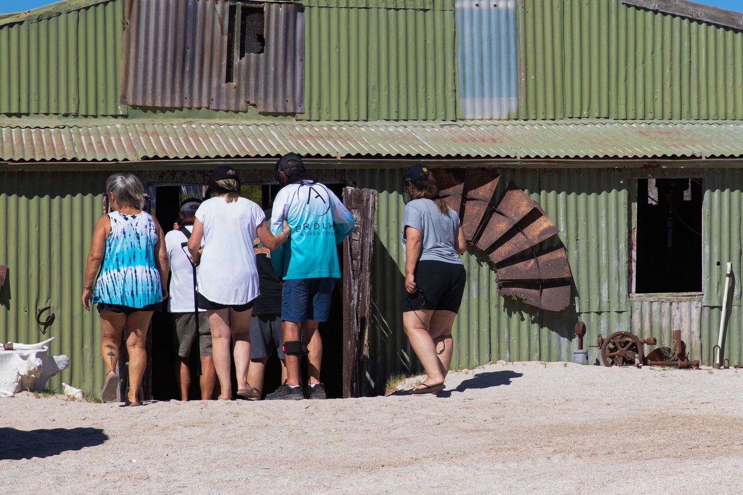 Group of tourists peering into a vintage green corrugated iron shed with rusty machinery outside.