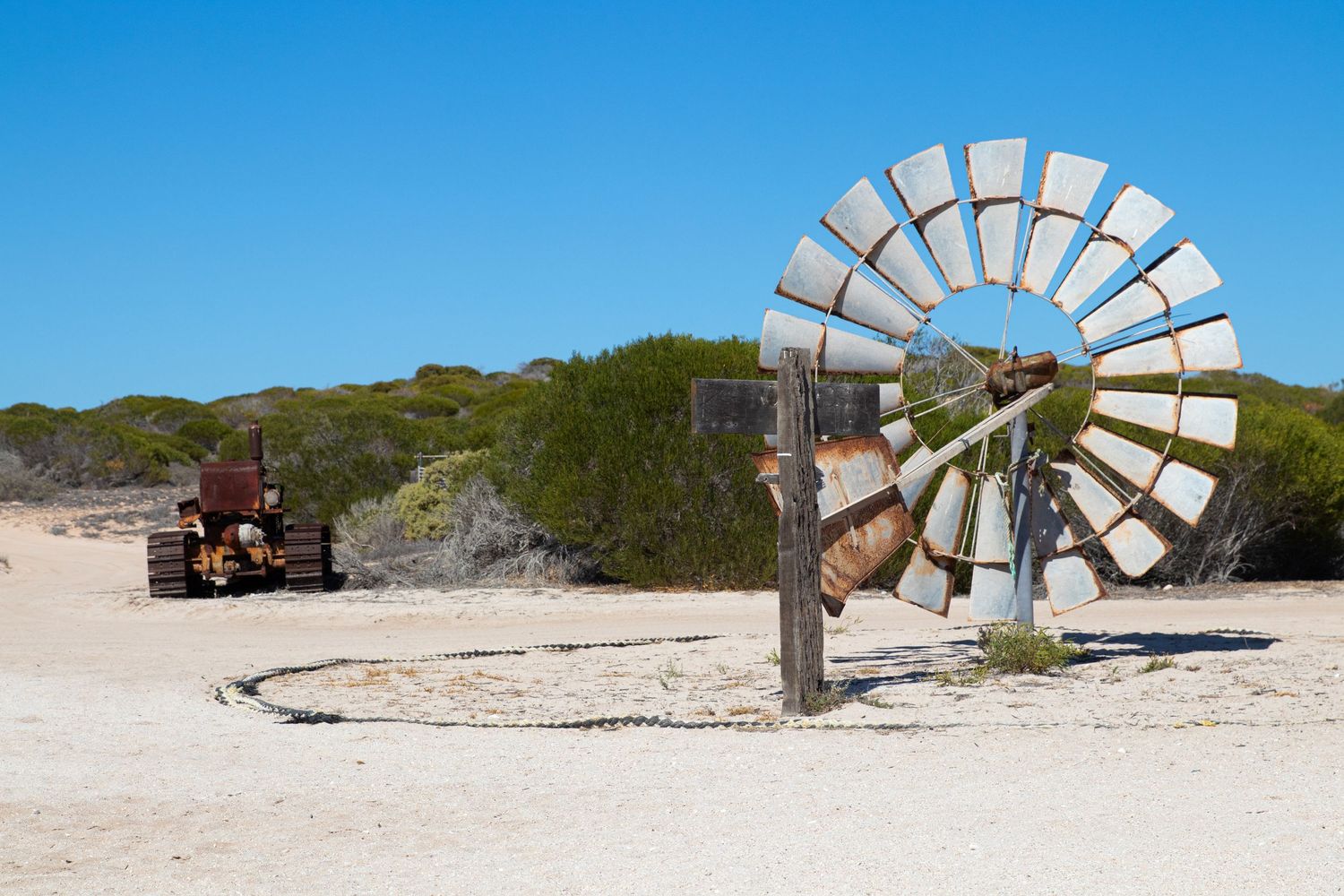 Rusty vintage windmill and old tractor abandoned in sandy desert landscape with blue sky and scrub bushes.