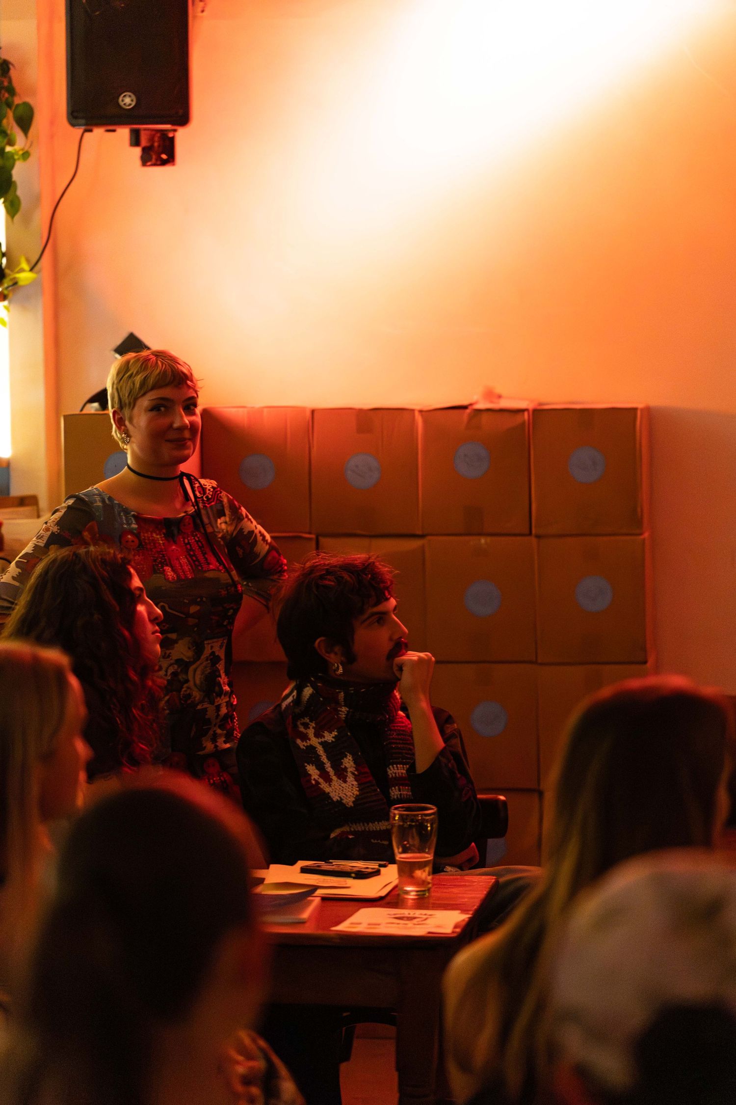 Two women in a warmly lit bar setting, one standing and smiling, the other seated at a table with a drink.