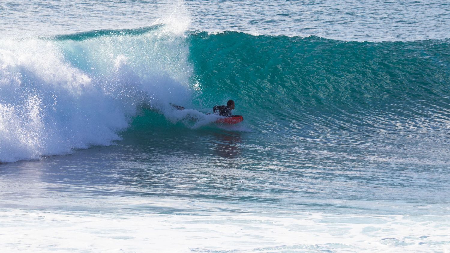 Bodyboarder in red suit tucked inside a perfect turquoise barrel wave in crystal clear ocean water.