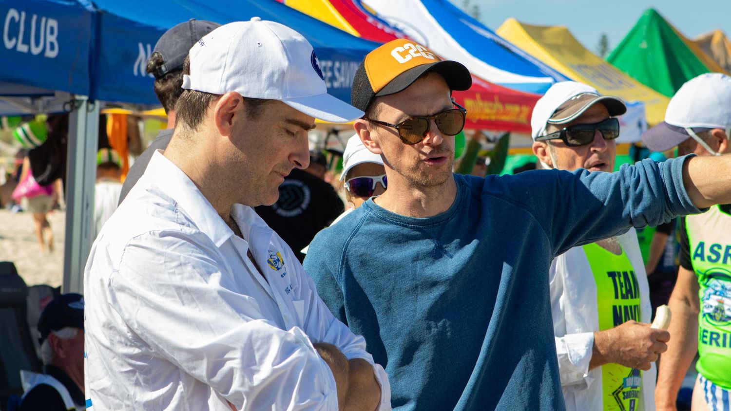 Two men shaking hands at a sunny beach surf lifesaving event with colorful tents in the background.
