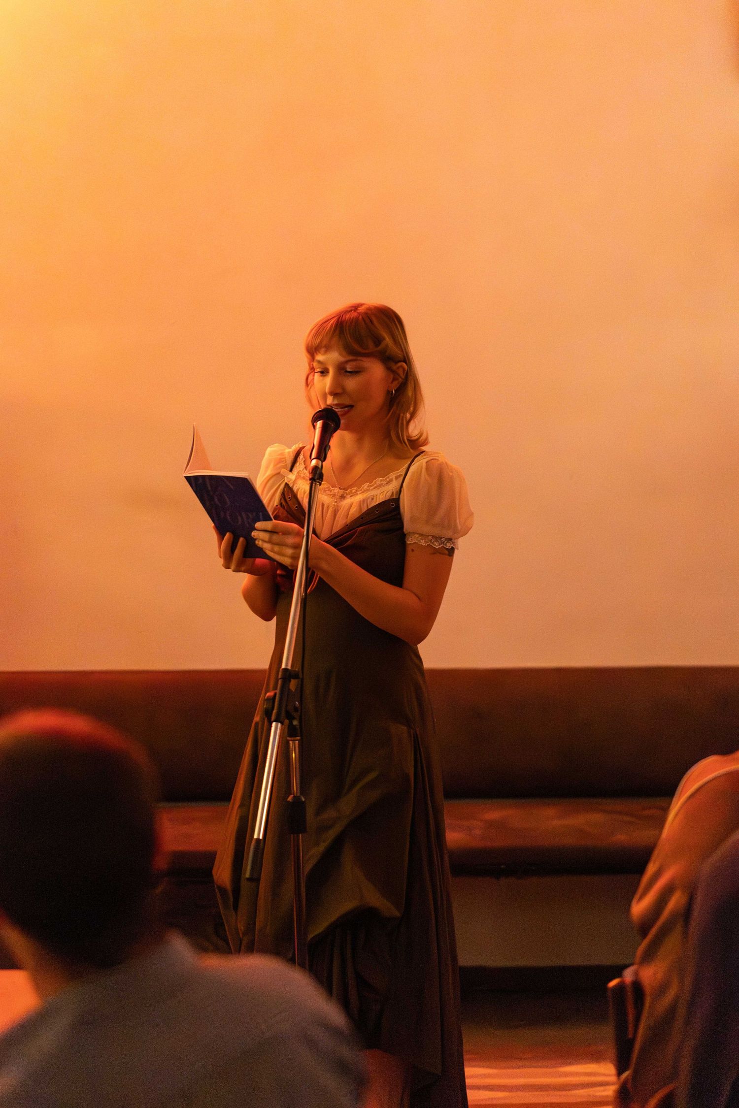 Young woman in a dark dress reads from a blue book at a microphone stand on a warmly lit stage.