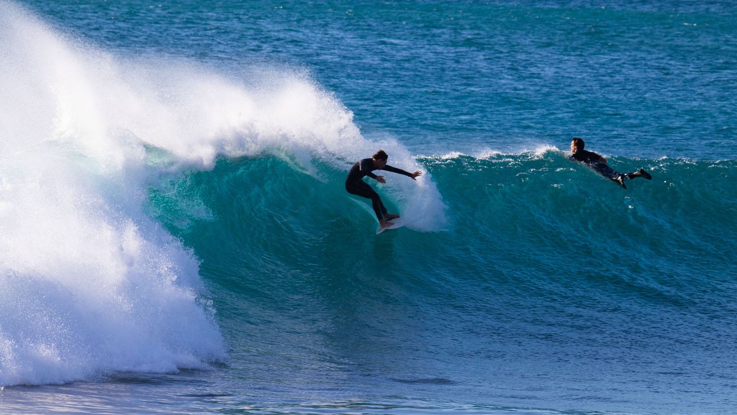Two surfers ride the same turquoise wave simultaneously, with one carving and one paddling nearby.