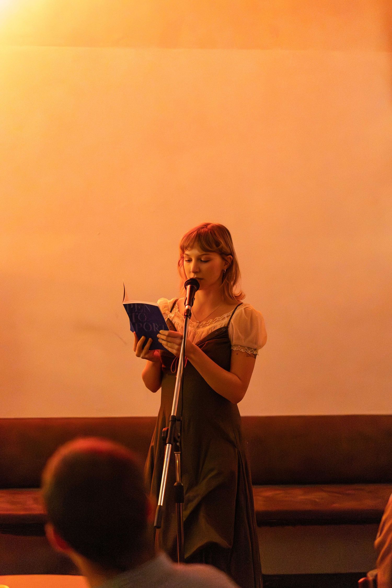Young woman in dark dress reads from blue book at standing microphone during spoken word event.