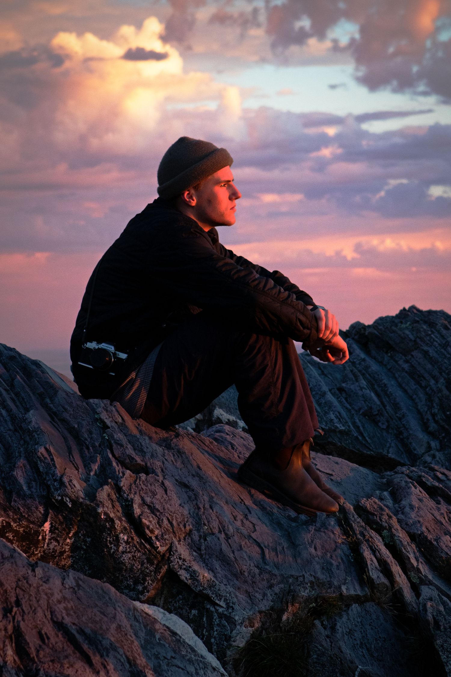 Man sitting on rocky mountain peak gazing into the distance beneath a dramatic pink and purple sunset sky.