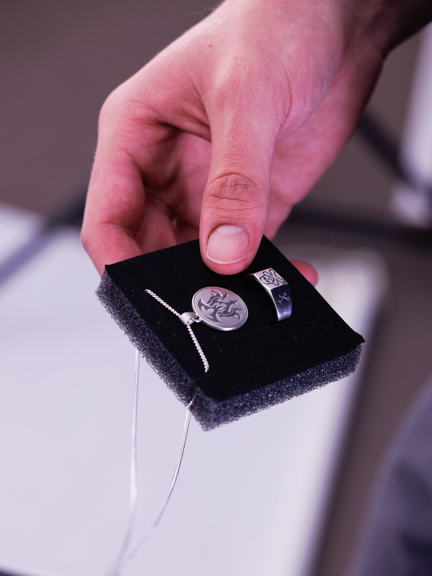 Hand holding a black foam jewelry display with a silver pendant necklace and engraved ring.