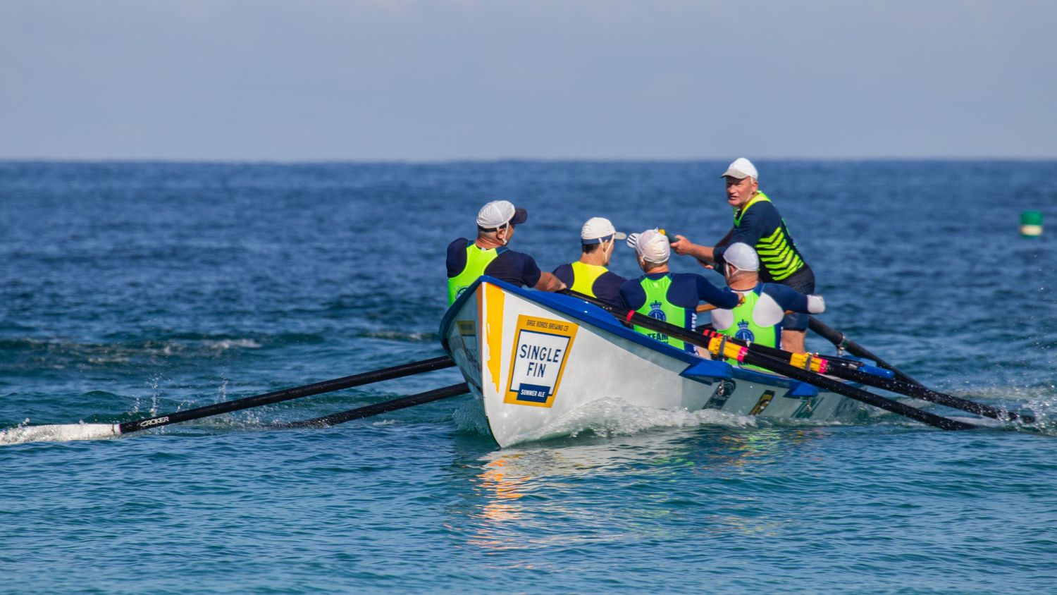 A team of rowers in yellow vests and white helmets power through ocean waves in a surf boat race.