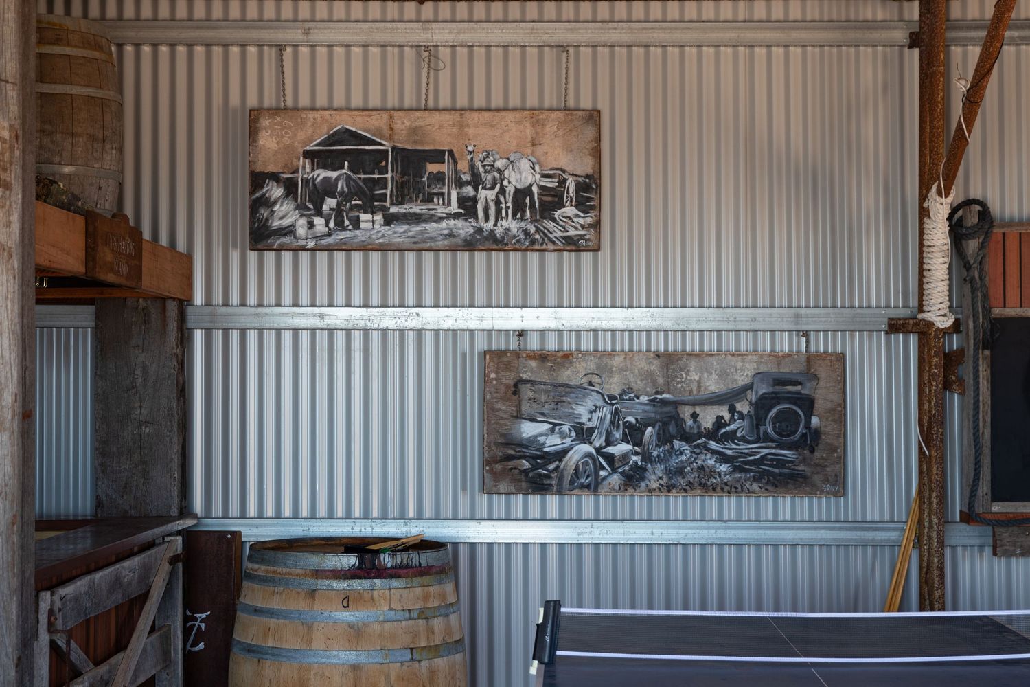 Vintage black and white artwork panels displayed on corrugated iron wall above a wooden barrel indoors.