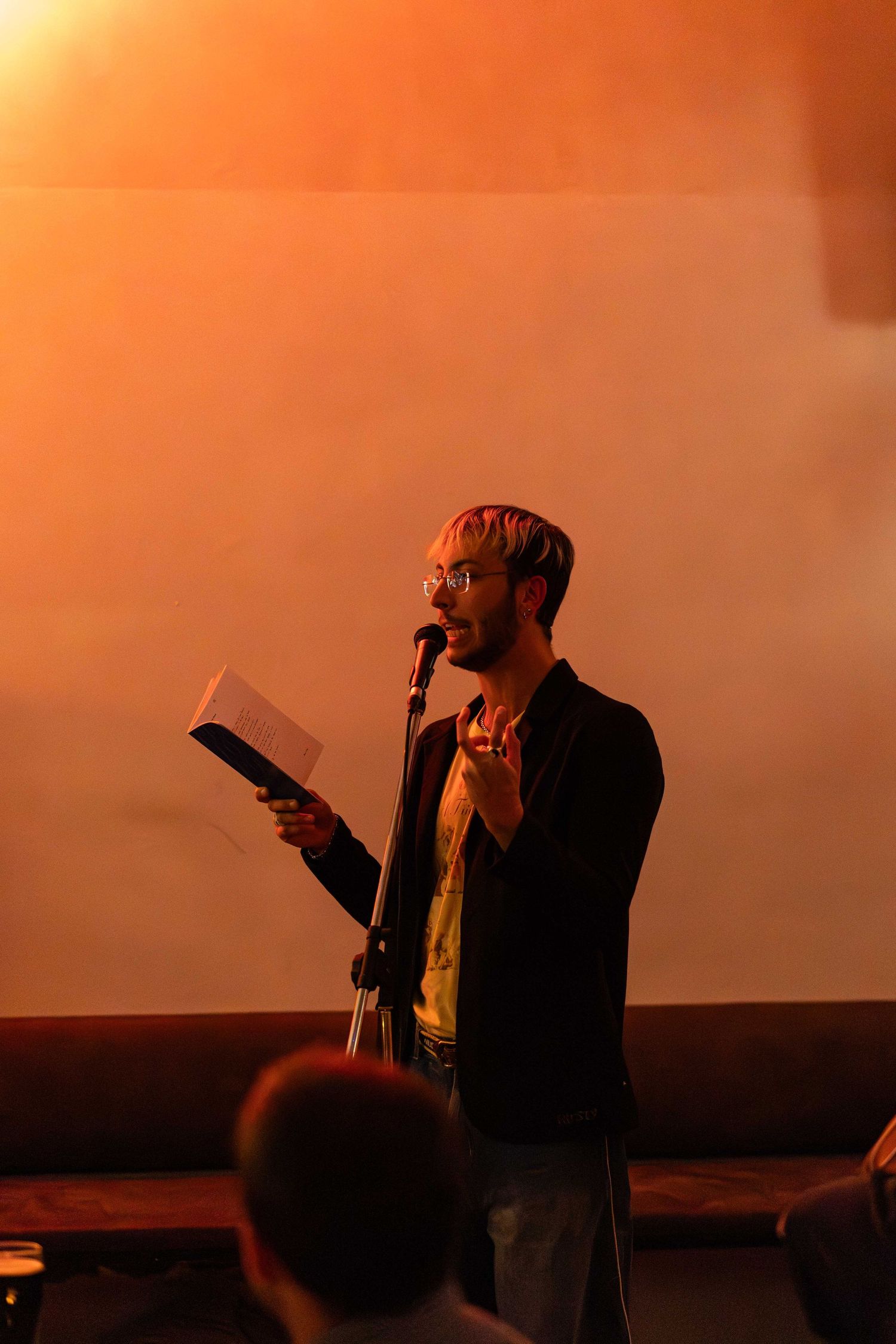 Man with glasses reads from paper at microphone stand under warm orange stage lighting.