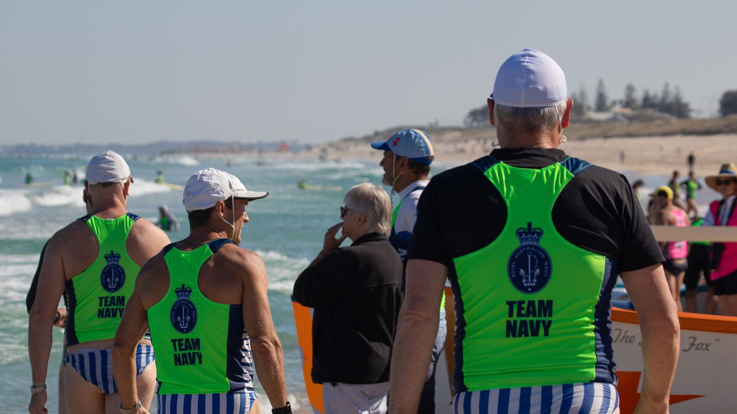 Three Team Navy surf lifesavers in green vests watch ocean activity at an Australian beach carnival.