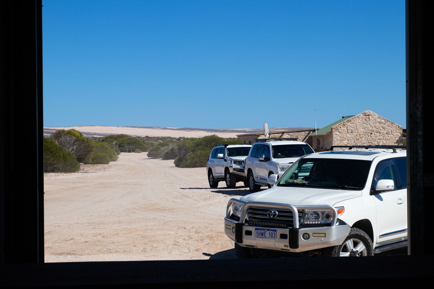 White Toyota 4WD vehicles parked on sandy ground near a stone building framed by a dark window border.