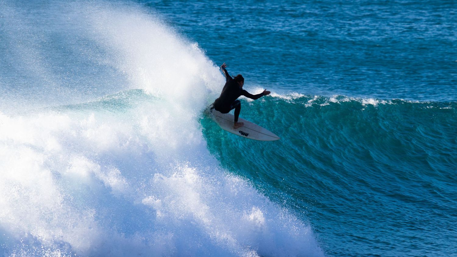 A surfer in a black wetsuit rides the crest of a massive turquoise ocean wave.