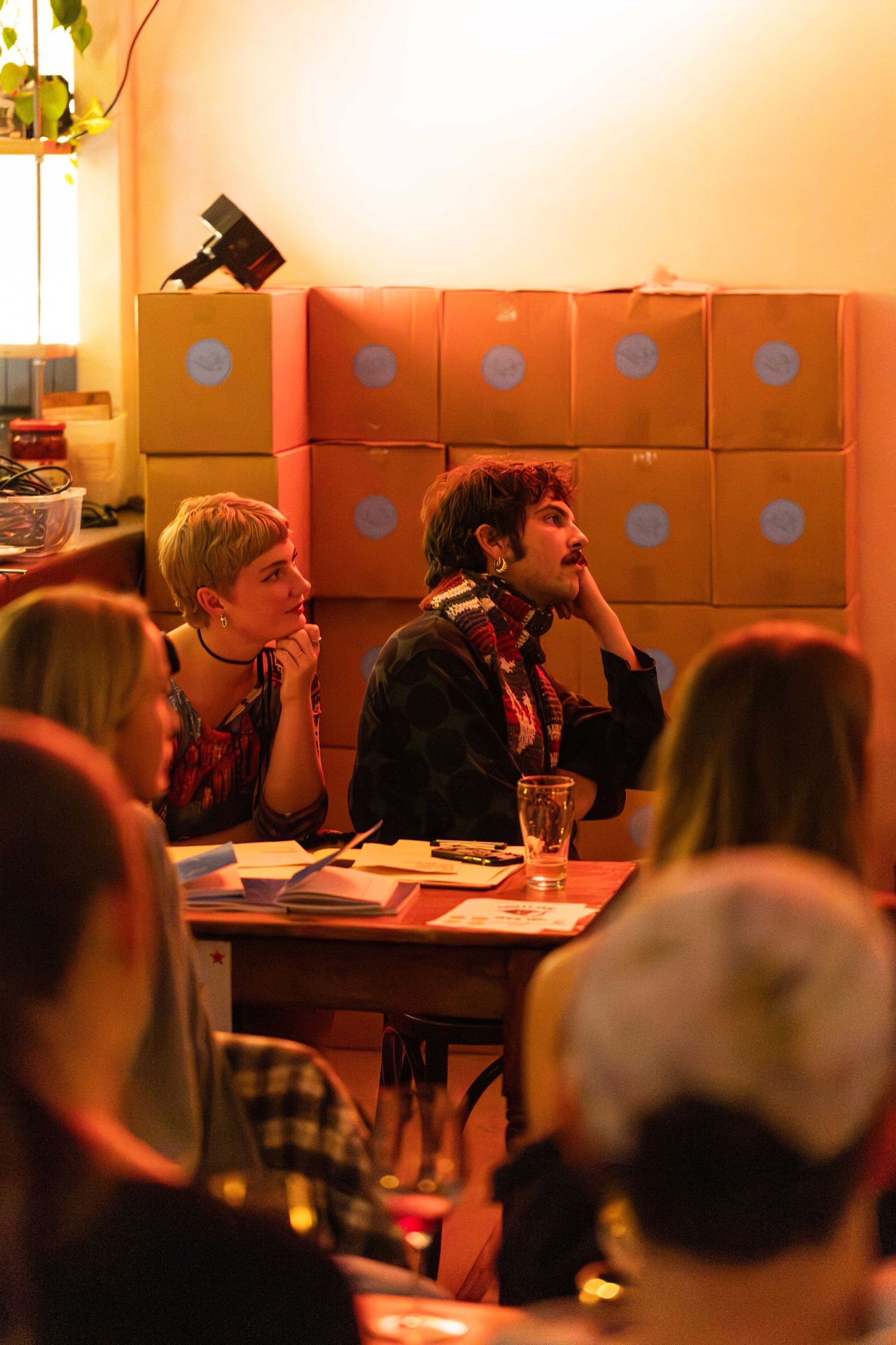 Two young people listening attentively at a table during a cozy indoor event with stacked boxes behind them.