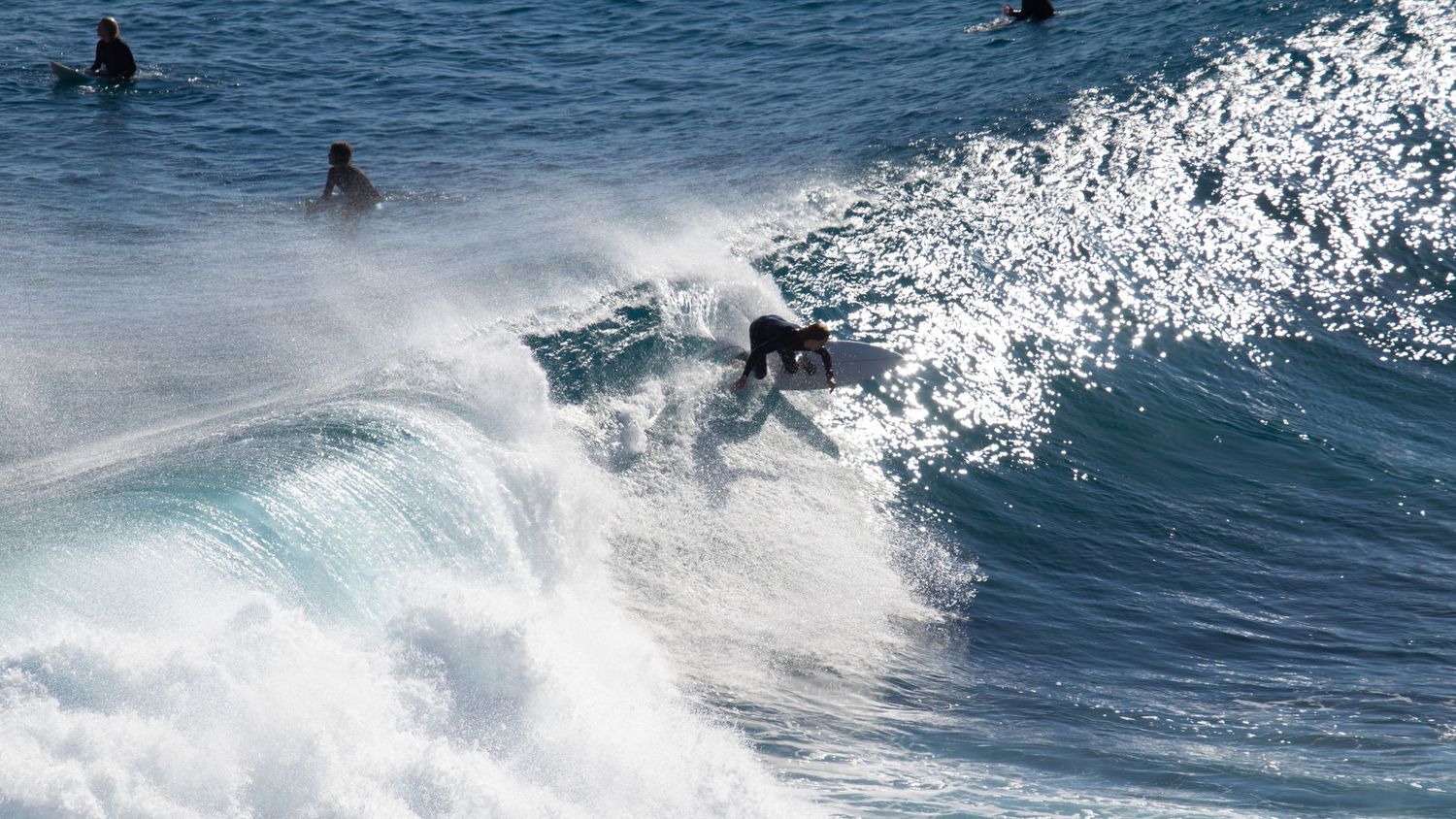 A surfer rides a foamy white wave close to shore while other surfers wait in the background.