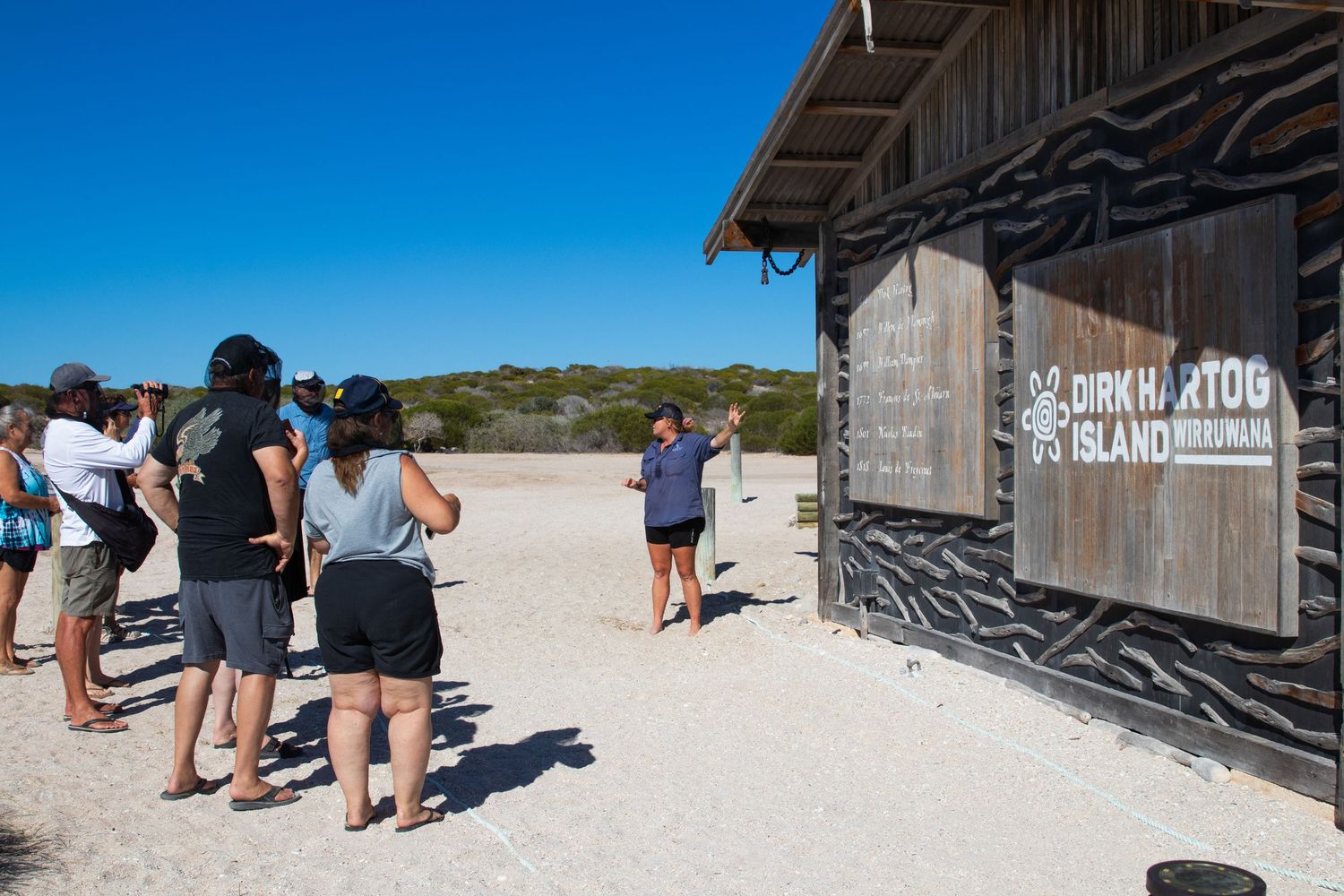 Tour guide addressing visitors outside Dirk Hartog Island Outstation rustic wooden building on beach.