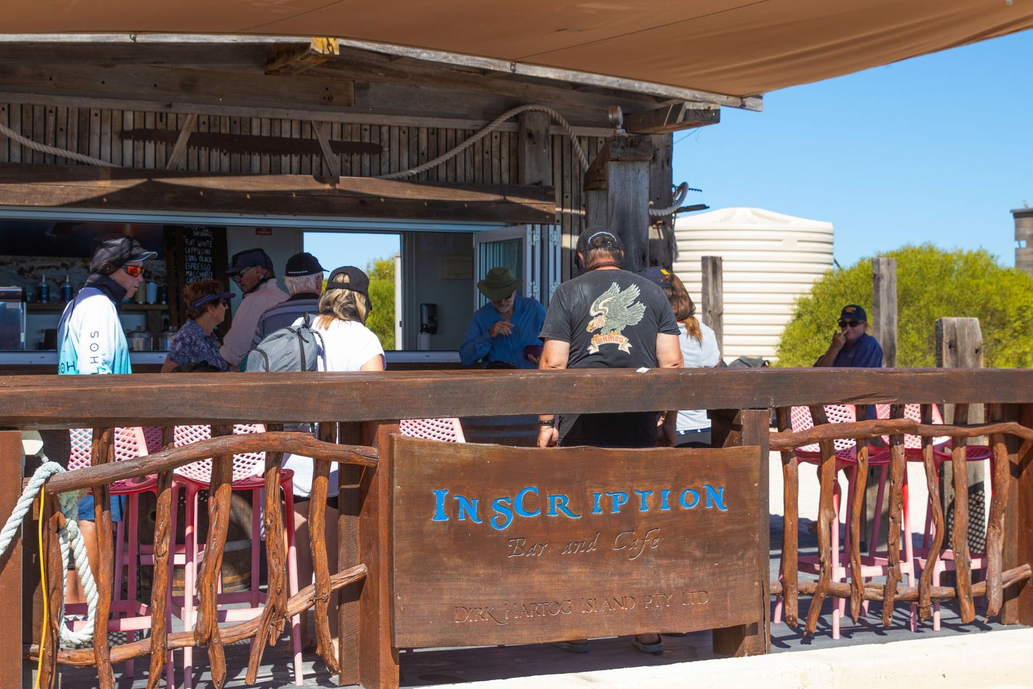 Guests gathered at the rustic Inscription Gin outdoor bar under a shaded structure on a clear day.