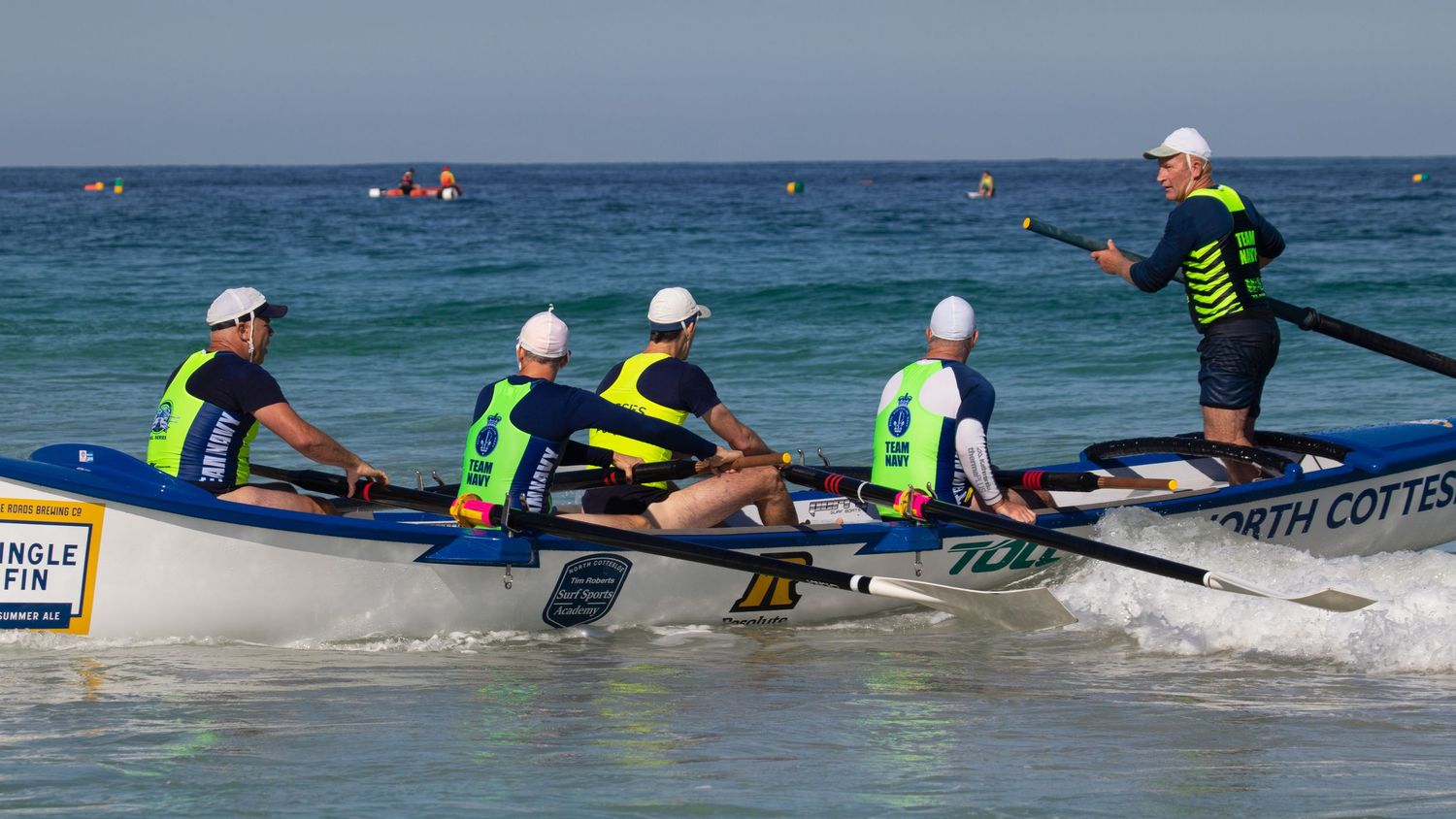 North Cottesloe surf boat crew rows through shallow waves with a standing sweep oarsman guiding them.