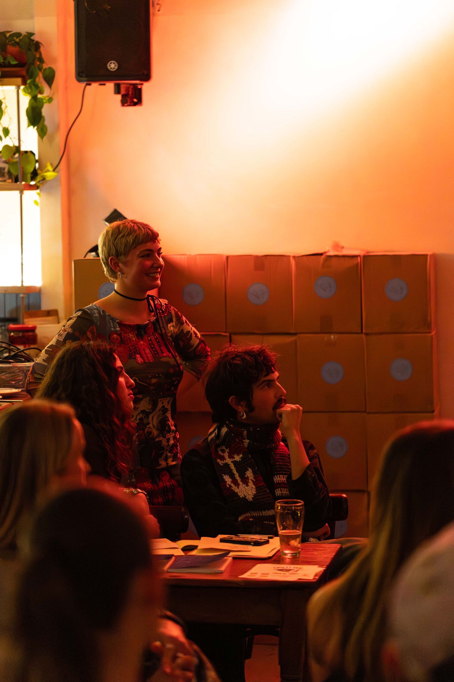 Two people seated near stacked cardboard boxes in warm orange-lit venue during a live event.
