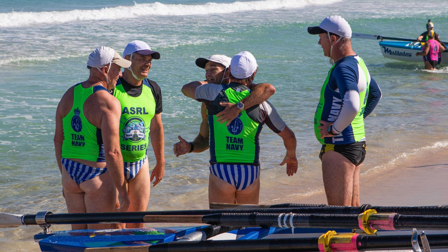 Surf boat rowing team in ASRL Series uniforms discuss strategy on a sunny Australian beach.