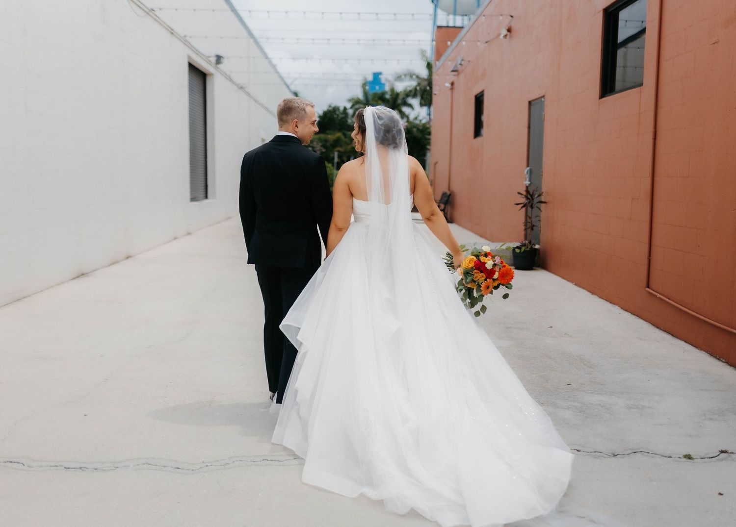 A bride and groom walk together down an outdoor corridor with coral-colored walls.