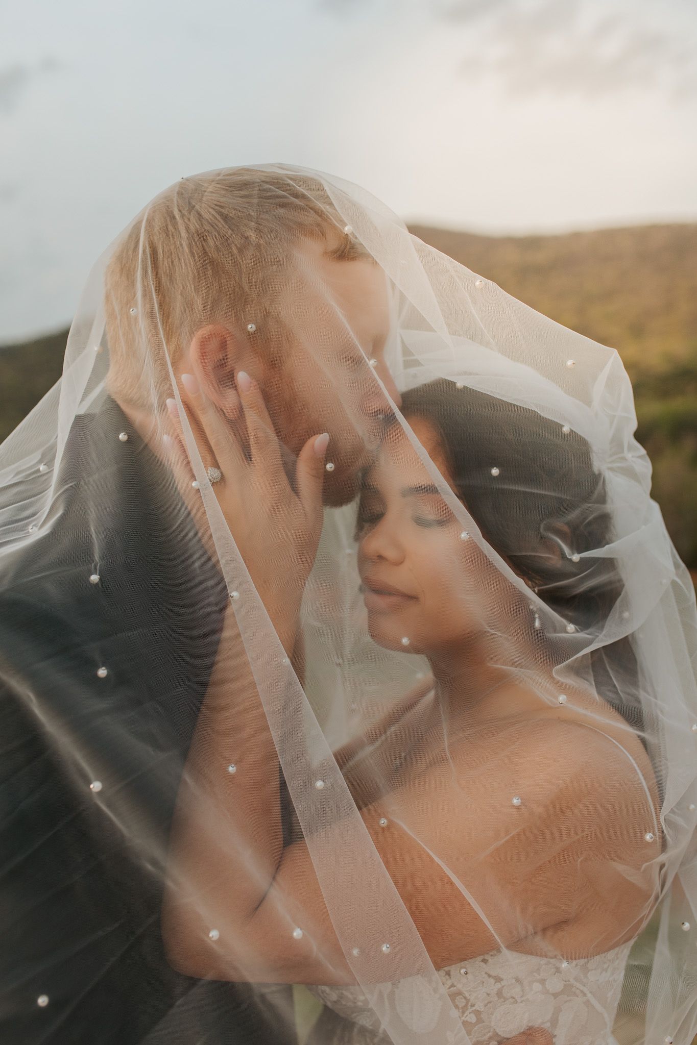 A romantic wedding photo shows a couple under a delicate veil sharing an intimate moment against a scenic outdoor backdrop.