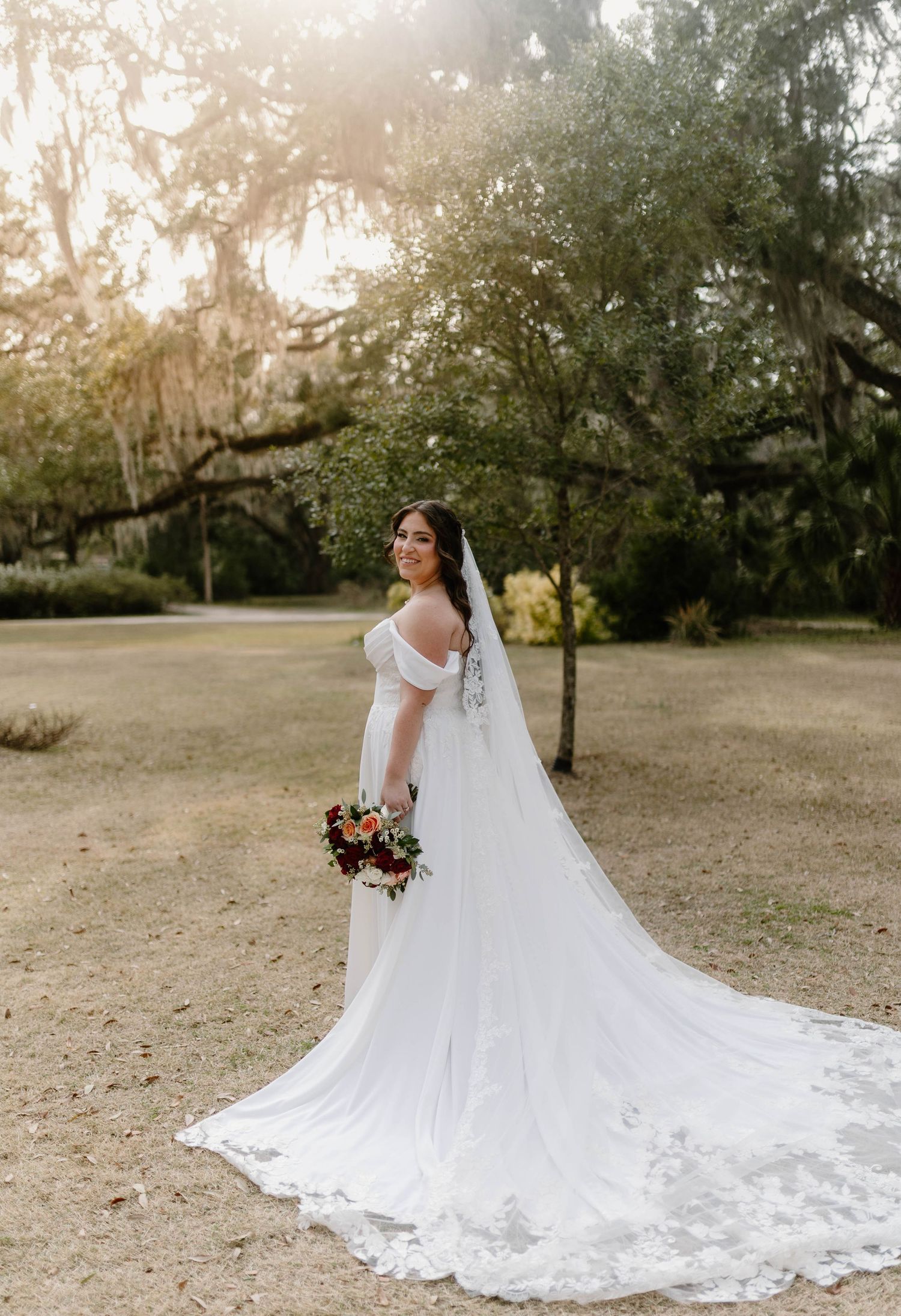 Bride in off-shoulder white gown with cathedral veil holds bouquet under Spanish moss trees at golden hour.