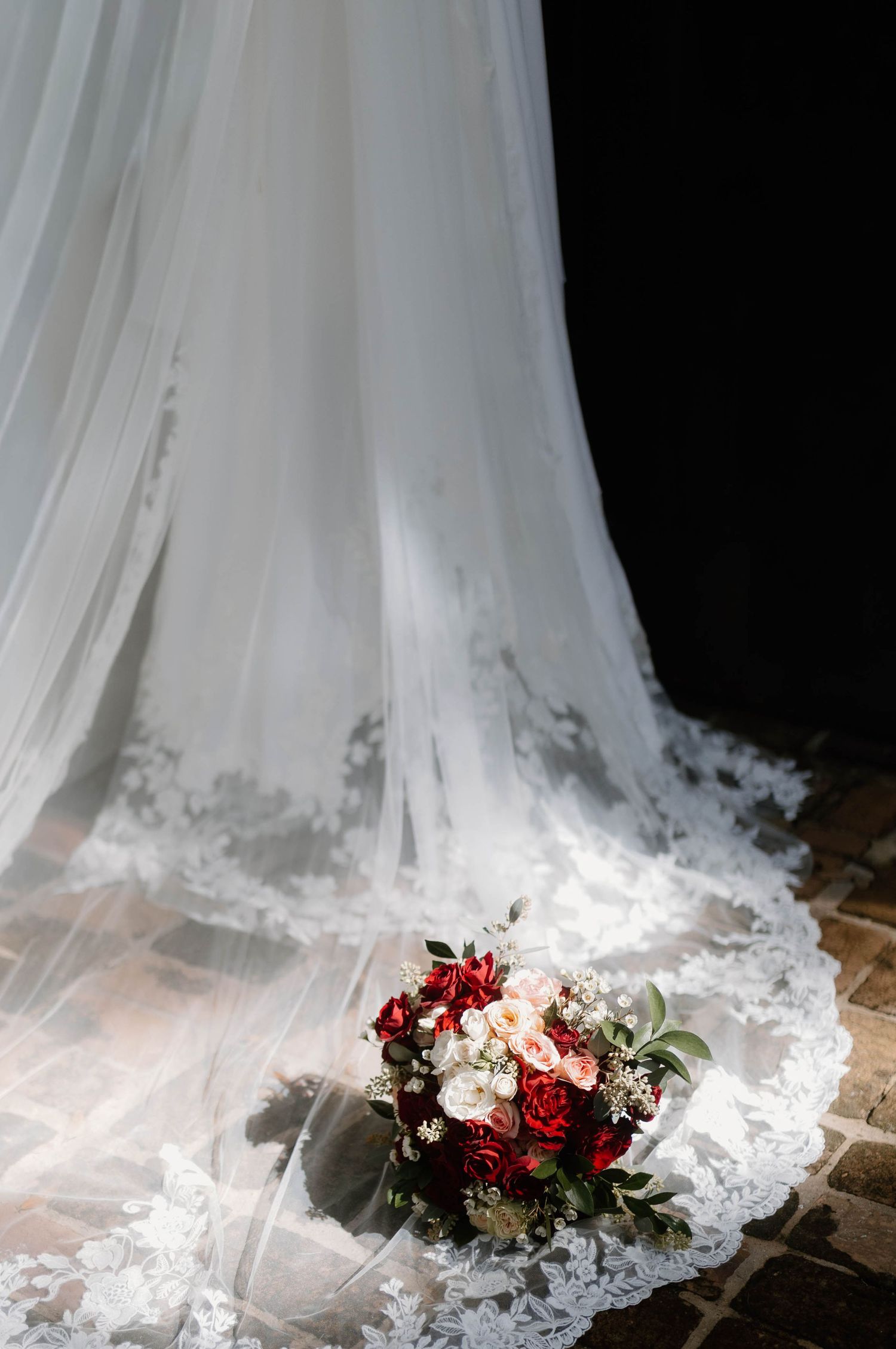 Bridal bouquet of red and white roses resting on lace wedding dress train on cobblestone floor.