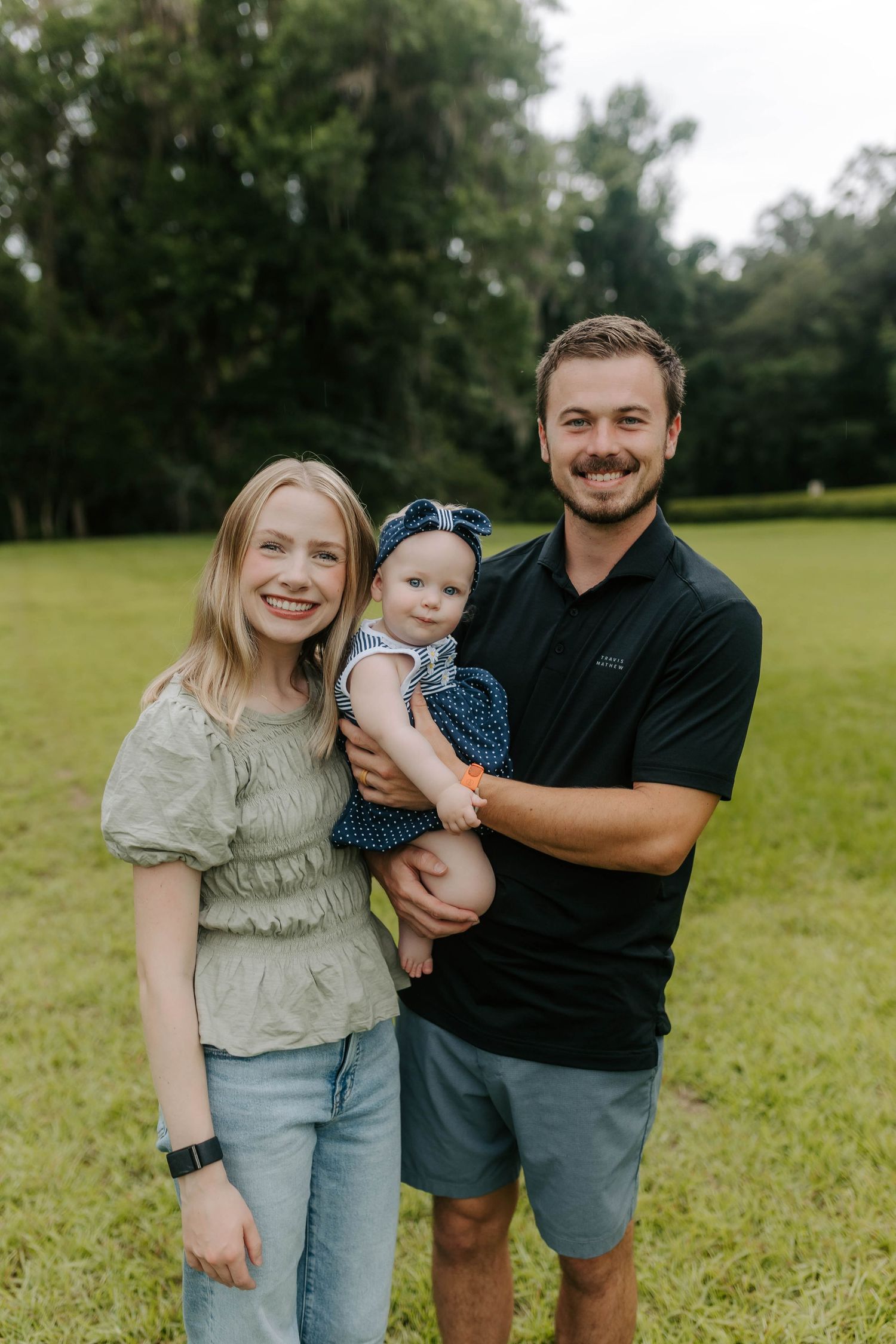 A young couple smiles outdoors in a green park while holding their baby girl in a blue polka dot outfit.