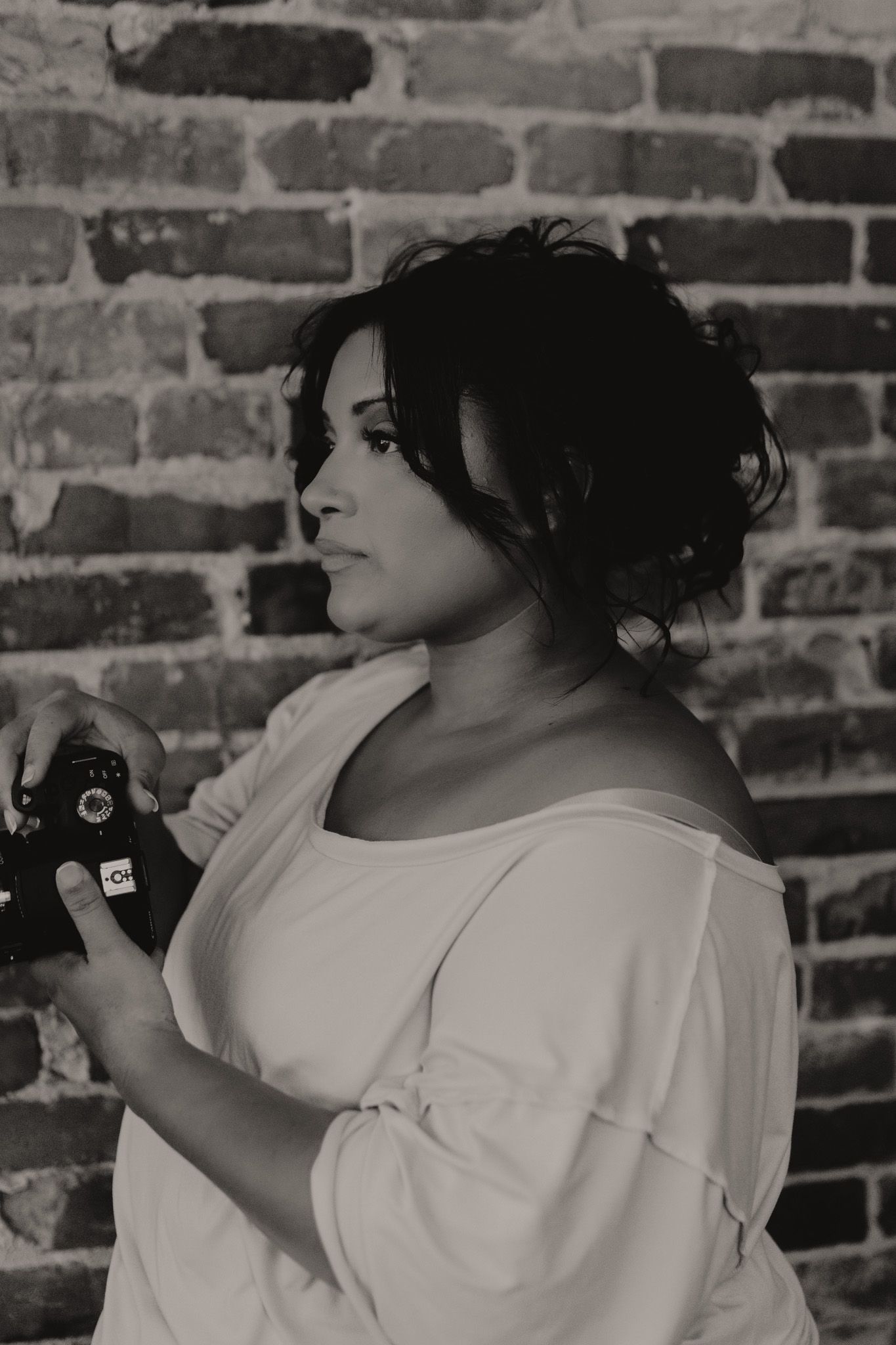 Photographer in white top poses with camera against brick wall background.