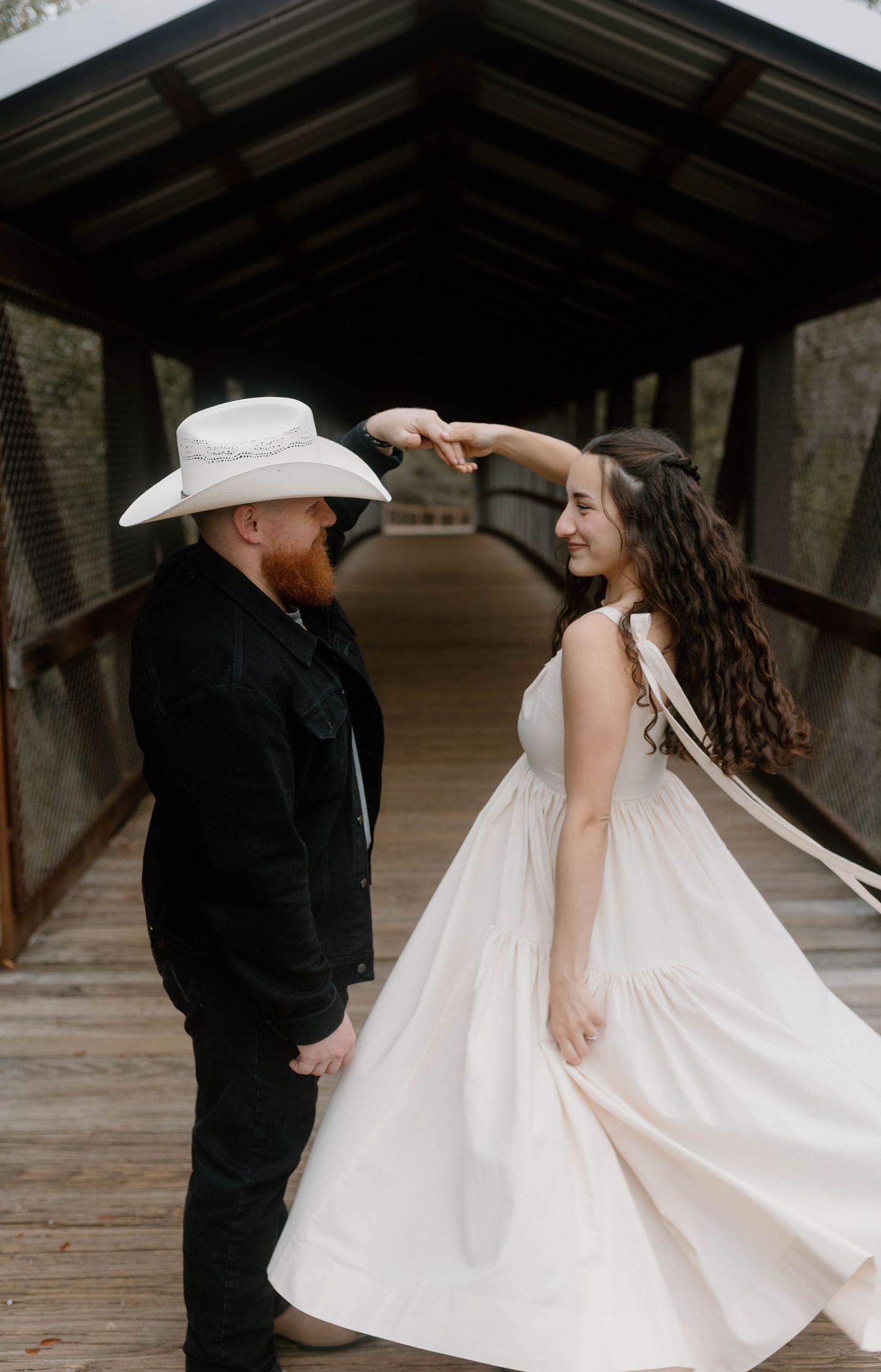 A couple dances on a covered bridge, man in black suit and white cowboy hat, woman in flowing white dress.