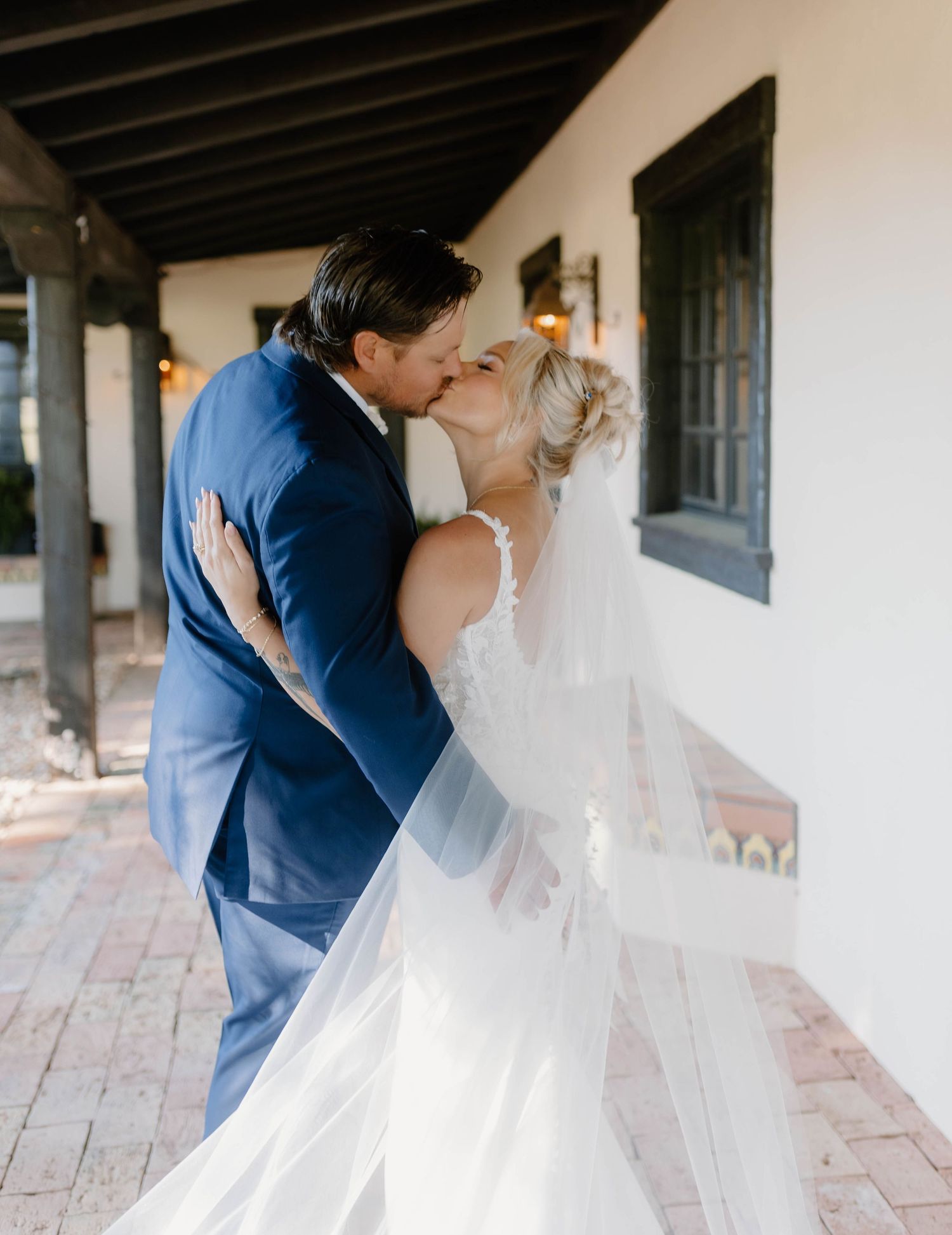 A bride and groom share a romantic kiss outside a white building, with the bride wearing a flowing veil and lace gown.